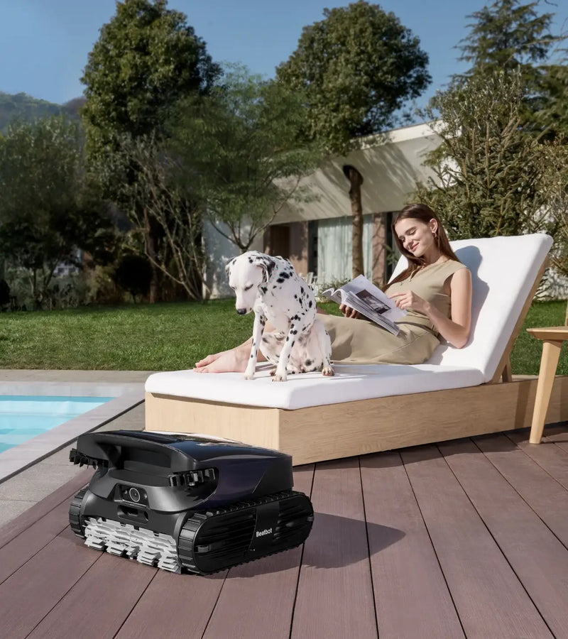 Woman with a dog on a lounge chair next to an AquaSense 2 Ultra robot pool cleaner on a wooden deck.
